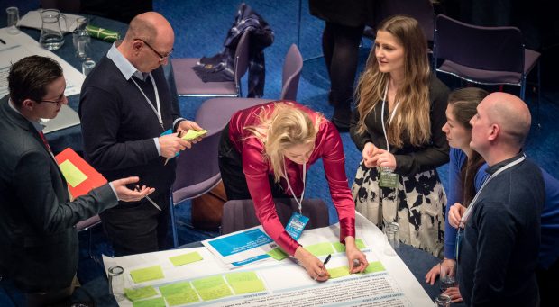 A working session at Transforming Together, a group of six people gathered round an A0 poster with post it notes covering the bottom of the sheet