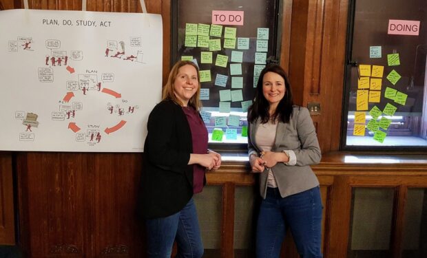 Two people - including the author, Rebecca Herbert - are standing in front of a wall. On the left of the wall is a poster titled 'plan, do, study, act' - in the middle are post its under a heading 'To Do' - and on the right are post its under the heading 'Doing'